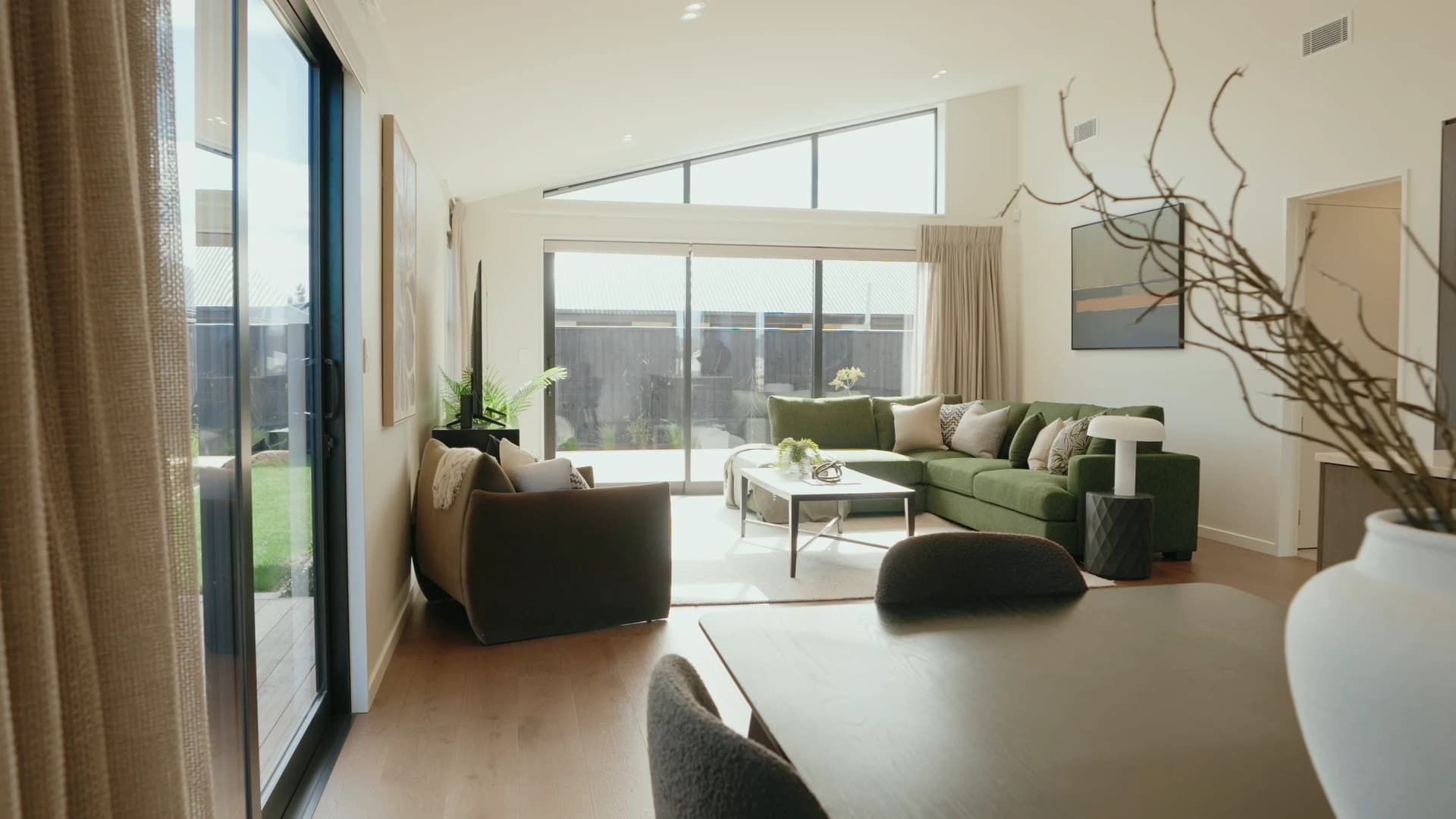 A living room in a Pringle home, a green corner sofa and a low table on a pale rug, sliding doors the width of the wall onto a courtyard, under a raked ceiling with a triangular clerestory window above