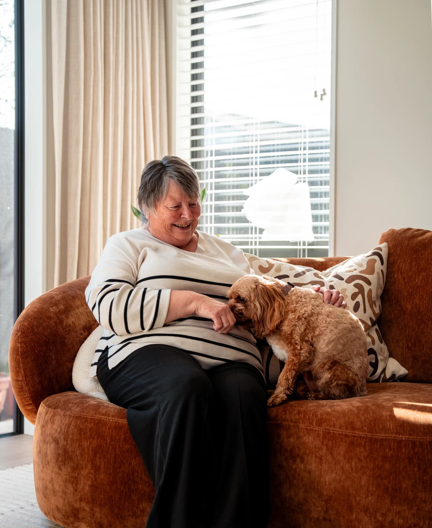 A woman in a striped jumper laughing on a rust velvet sofa as a small curly-haired dog noses at her hand, sheer curtains and a window with the blinds up behind her