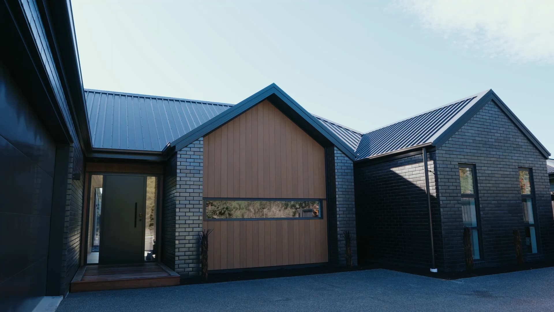 A Pringle home from the drive: a cedar-clad gable with a slot window between two dark brick wings under a steel roof, in low sun
