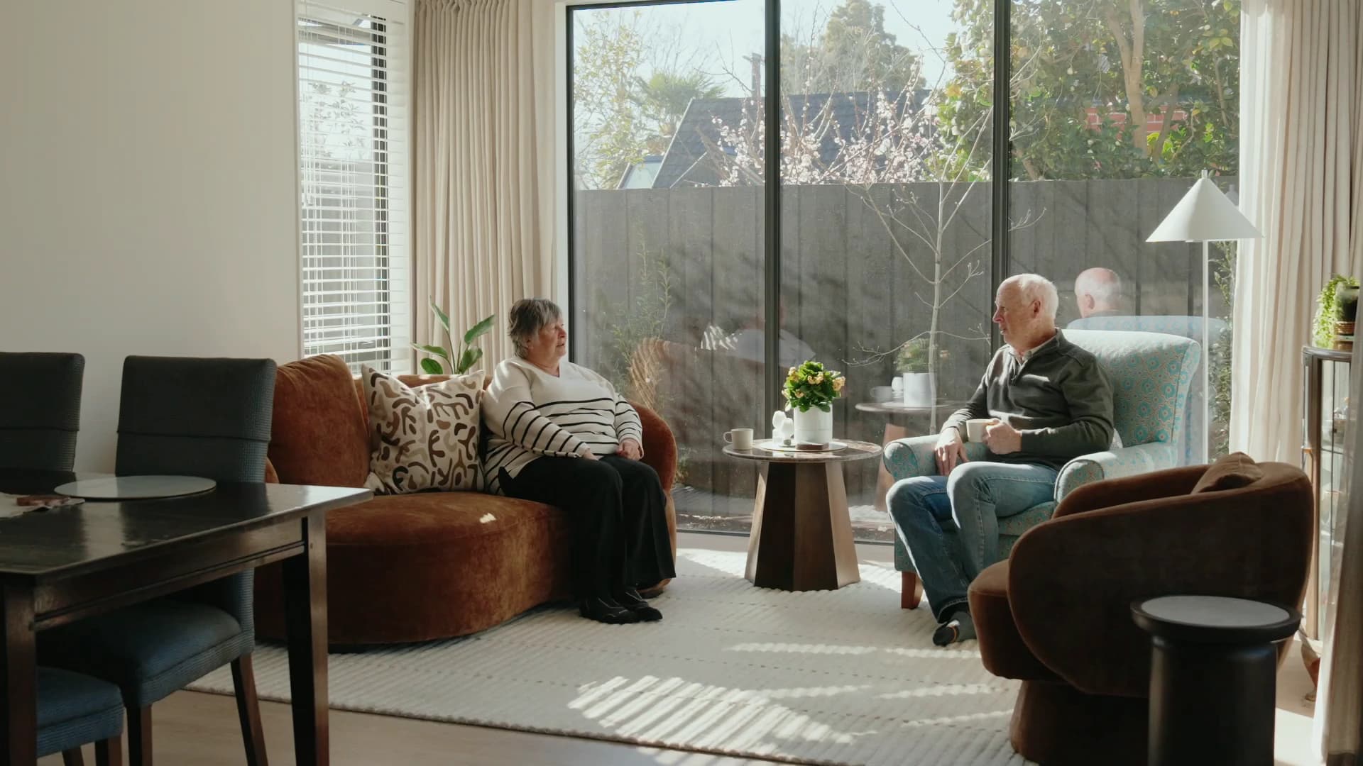 A living room in low sun, full height sliders onto a walled courtyard, a rust sofa and a patterned armchair either side of a low table, two people sitting with cups of tea