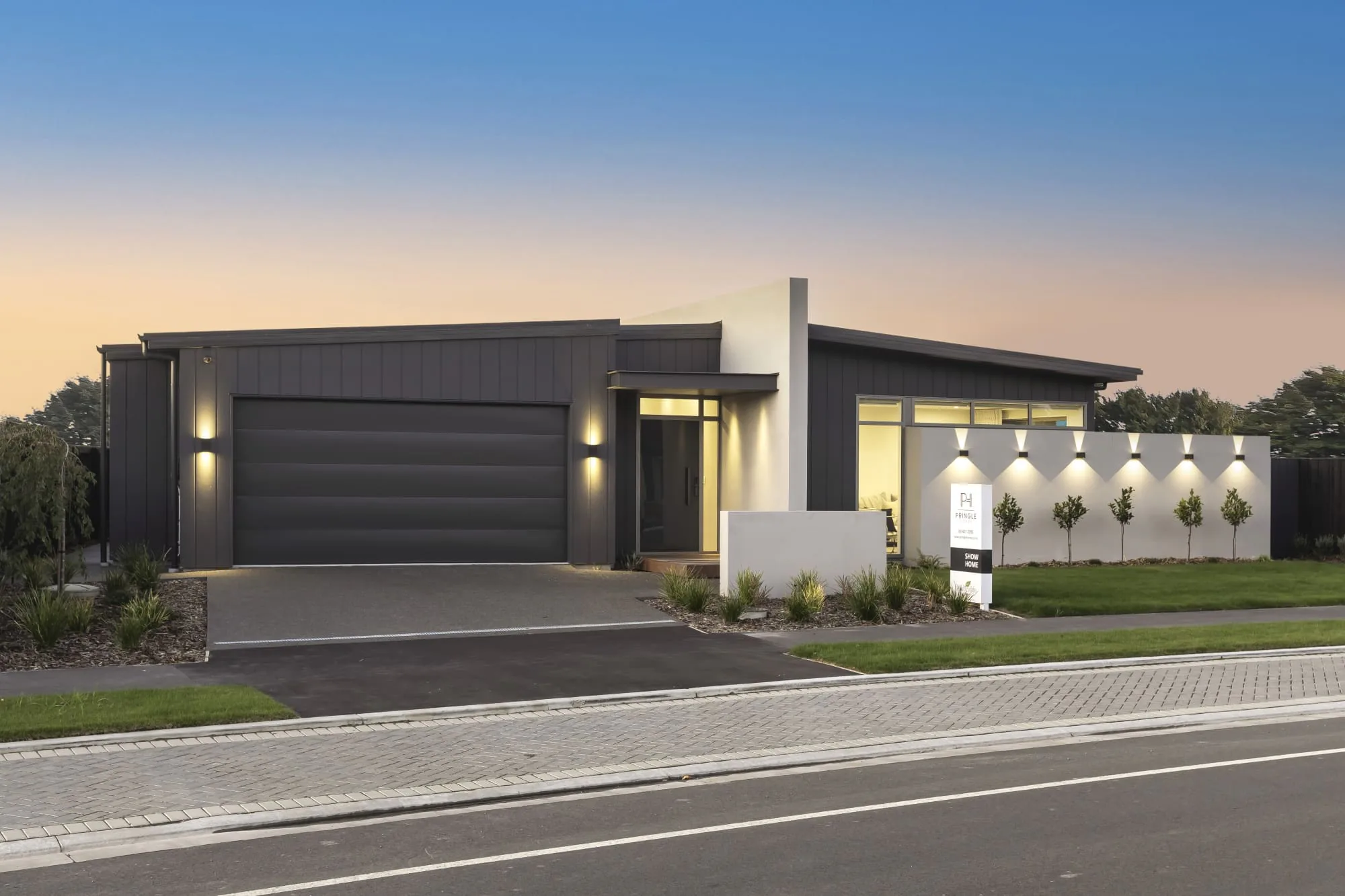 A Pringle Homes show home at dusk, dark cladding and lit windows seen from across the street