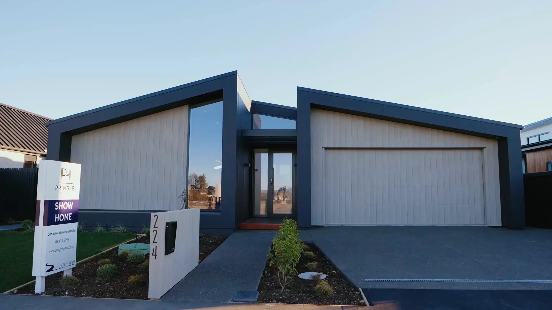 A Pringle show home at Meadow Stream from the street: pale vertical cladding framed in dark trim under two skillion roofs, with a Pringle show home sign on the lawn